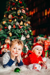 Fototapeta na wymiar Two pretty little boys lie on fluffy carpet before a Christmas tree