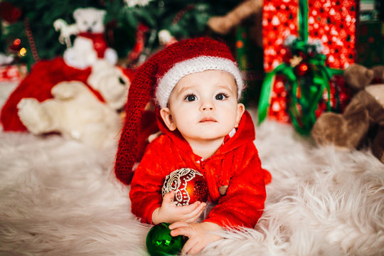 Pretty little boy in red suit lies on fluffy carpet before a Christmas tree