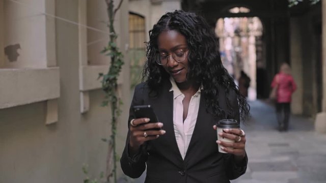 Beautiful young afro american black woman with long black hair and glasses,wears casual professional business outfit on way to work or meeting, chats or checks schedule on smartphone with coffee to go