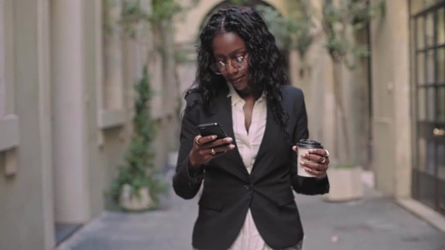 Natural afro american beauty business woman in stylish outfit for office work, walks on street with take away coffee cup,looks at screen of smartphone,checks emails and notifications from applications