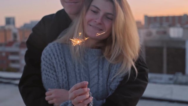 Slow motion shot of loving and caring young couple in 20s, hug, kiss and cuddle on top of rooftop at sunset, burn sparklers to celebrate new years eve or just have fun on romantic date