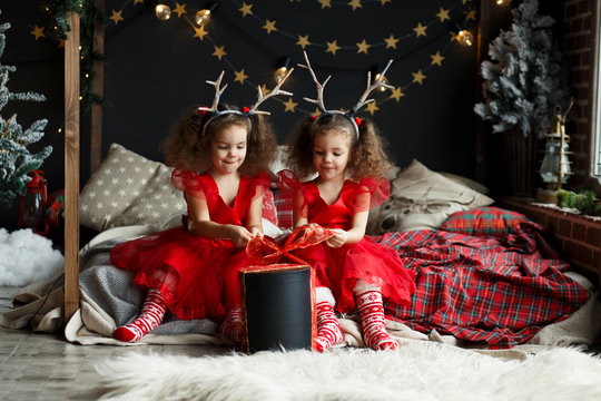 Children Opening Christmas Present. Two little todder girl with deer horns on head. Twins in red dresses sitting on the bed in cozy room.