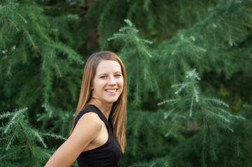 Naklejka premium Amazing Smiling Woman in Black Shirt with Long Shine Hair Standing near Spruce Tree During Summer Time.