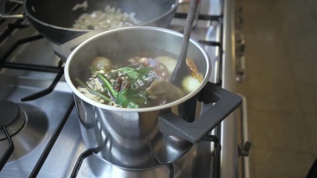 Slow motion shot of industrial kitchen stove with metal silver pot full of aromatic tasty and delicious soup, female chef hand stirs ingridients with wooden spoon, concept grandma's recipe