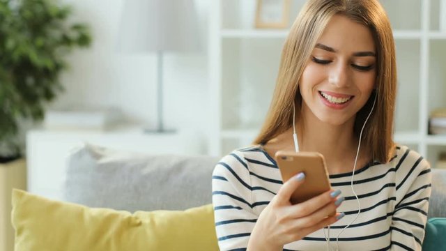 Attractive young woman listening to music in white headphones and using smart phone for chatting at home. Close up.