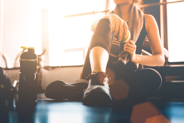 Sport woman sitting and resting after workout or exercise in fitness gym with protein shake or drinking water on floor. Relax concept. Strength training and Body build up theme. Warm and cool tone - obrazy, fototapety, plakaty