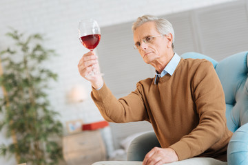 Wine expert. Low angle shot of a serious elderly man sitting in a chair and concentrating on a glass of red wine while examining it at home.