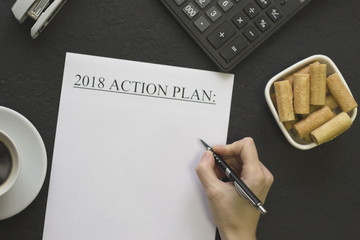 Hand writind 2018 action plan, a white bowl of cookies, coffee and office supplies on a black concrete background, top view