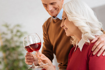 Cheers. Selective focus on a harmonious elderly couple hugging and smiling while enjoying their glasses full of red wine.