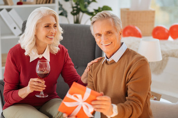 He is my one and only. Selective focus on a relaxed senior lady looking at her husband with a cheerful smile on her face while both enjoying their glasses of wine and exchanging present at home.