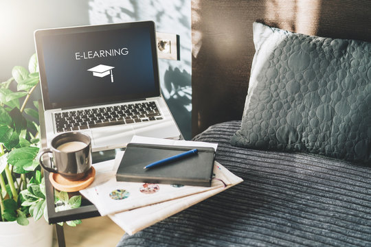 Bedroom,workplace without people,close-up of laptop with an inscription- e-learning on screen on table, desk. Nearby is newspaper, cup of coffee, notebook. Nobody. Online education, distance learning.