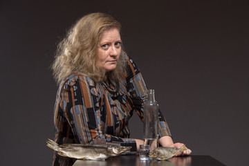 Lonely mature fat woman alcoholic drinks vodka from glasses and wine glasses and snack cucumber and fish