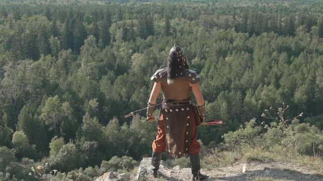 Mongolian archer stands on edge of mountain on sunny day. Young man dressed in clothes of traditional culture, holds bow and arrow and looks around space opened front of him: mighty green forests
