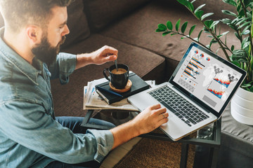 Rear view. Young man, entrepreneur, sits at home on couch at coffee table,drinking coffee,working on laptop with graphs, charts, diagrams on screen. Online marketing, education, e-learning. Startup. - obrazy, fototapety, plakaty