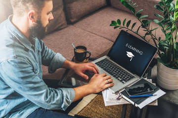 Young bearded hipster man,entrepreneur sits on couch at coffee table,uses laptop with inscription- e-learning on screen.On table notebook,cup of coffee.Online marketing,education,e-learning.Startup. - obrazy, fototapety, plakaty