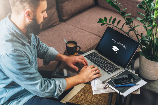 Young bearded hipster man,entrepreneur sits on couch at coffee table,uses laptop with inscription- e-learning on screen.On table notebook,cup of coffee.Online marketing,education,e-learning.Startup.