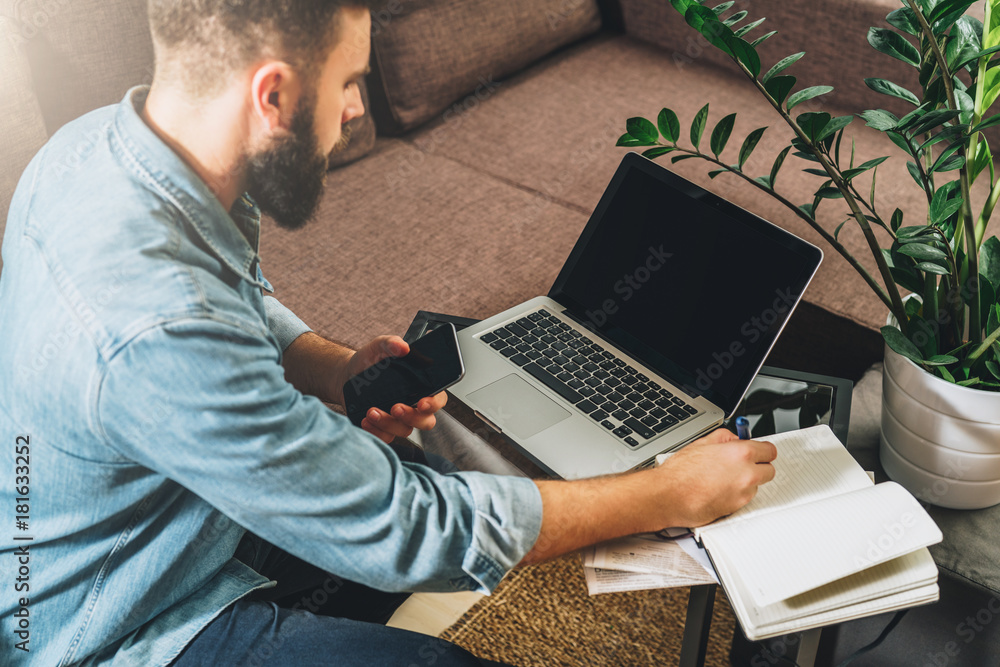 Wall mural Young bearded man sits at table, holds smartphone in his hand, uses laptop and makes notes in his notebook.Guy working, studying.Online marketing, education, e-learning, e-commerce. Startup, planning.