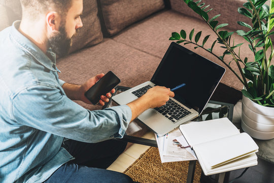 Rear view. Young bearded hipster man, entrepreneur, dressed in denim shirt, sits at home on couch at coffee table, holding smartphone, showing pencil on screen of laptop. Online marketing, e-learning.