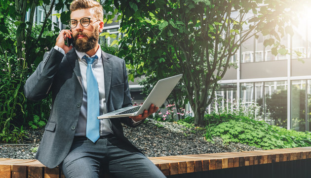 Summer day. Young bearded businessman in suit and tie sitting in park on bench, holding laptop and talking on cell phone. Man is working, studying online. Online marketing, education, e-learning.