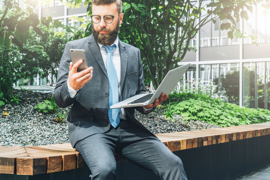 Summer day. Young bearded businessman in suit and tie sitting in park on bench, holding laptop and using smartphone. Man is working, studying online. Online marketing, education, e-learning.
