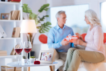 She said yes again. Selective focus on a table with two glasses of red wine and a velvet ring box with a retired couple exchanging gifts in the background.
