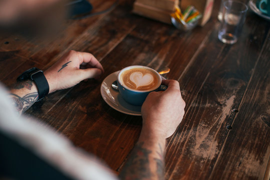 View of hipster man with authentic artisan tattoo hold cup of freshly brewed coffee with latte art heart made by barista, in blue cup at local cafe or bar, sits behind wooden aged table
