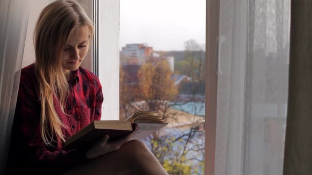 Beautiful young blonde woman reading a book and sitting on the windowsill