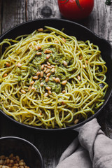 Close-up of Italian spaghetti with pesto sauce and pine nuts in the black cast-iron frying pan on the rustic wooden table.