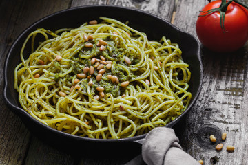 Close-up of Italian spaghetti with pesto sauce and pine nuts in the black cast-iron frying pan on the rustic wooden table.