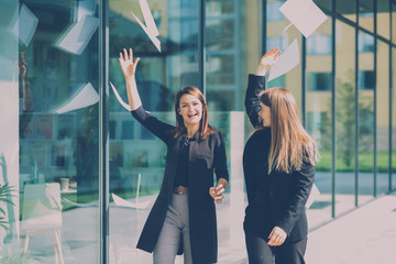 Two happy business woman throwing documents in the air while going along modern building. Freedom...