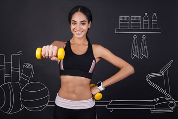 Fototapeta na wymiar Maintain healthy way of life. Cheerful content smiling woman holding dumbbells and doing sport exercises while standing in a gym