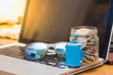 Travel and saving Concept. Close up of blue miniature baggage with sunglass and full of coins in clear bottle on laptop computer