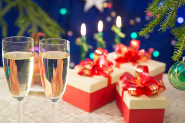 on the table glasses filled with champagne on a dark blue background with blurred lights. near there are New Year's gifts decorated with red bows.  a fragment of the New Year tree