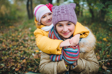 The younger sister hugs her older sister. Happy smiling children are having fun in the autumn park.