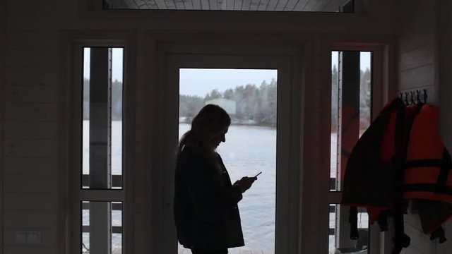 silhouette of a girl with a phone stands by the window with a lake view