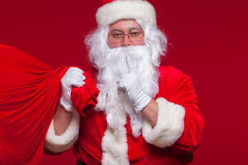 Portrait of Santa Claus with huge red sack keeping forefinger by his mouth and looking at camera