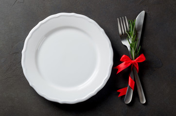 Festive set of cutlery knife and fork with red satin bow and rosemary, a white plate, dark stone slate background, top view, copyspace