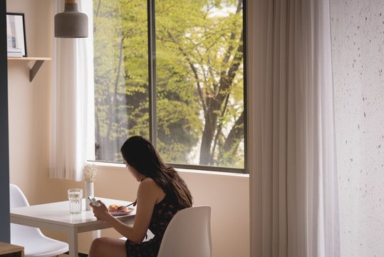 Side view of woman using phone while having breakfast at home