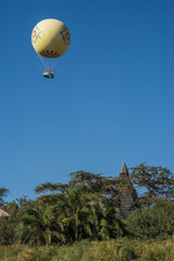Fototapeta na wymiar Large colorful air balloon with a gondola for several people hanging above a palm tree grove and a wooden triangular hut.