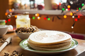 Christmas wafers placed on a wooden kitchen table, on a green plate next to a window. Wafers are round, cream color, surrounded by ingredients
