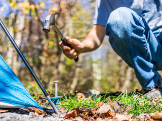 Young man setting up camp tent for camping trip on campground by hammering nail to the ground with hammer on corner