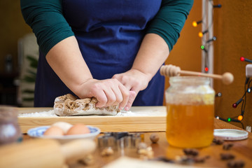 Close up of young woman hands kneading a dough on a wooden board. She is standing in the kitchen, behind a table, next to a window decorated with christmas lights