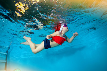 The little girl floats underwater in a cap of Santa Claus in the pool on a blue background. Portrait. Shooting under water. Horizontal orientation