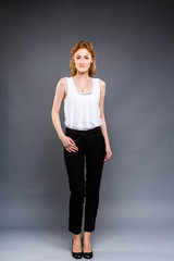 beautiful young Caucasian woman with long red hair in high heels, black trousers and a white shirt stands in full growth on a gray isolated background.