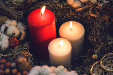 One red and two white burning candles in a wreath with natural christmas decorations