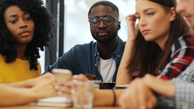 Cheerful group of young multiethnic friends in the city coffee shop drinking coffee in the morning. Close up shot of a pretty caucasian girl using a smartphone.