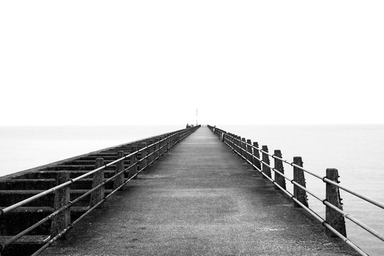 black and white photograph of a concrete pier with railings on each side converging into a vanishing point on the horizon, calm sea on either side of the pier