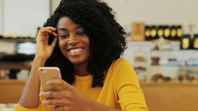 Close up portrait of the beautiful afro-american girl holding her mobile, surfing the Internet and texting her friends in the vintage cafe. Indoor.