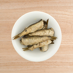 Top view of canned smoked sprats in a small white bowl atop a wood table.