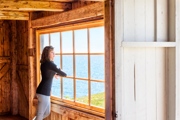 Fototapeta na wymiar Side of one, lonely, alone young woman standing by large glass window looking at peaceful ocean view, cliff, in rustic wooden house